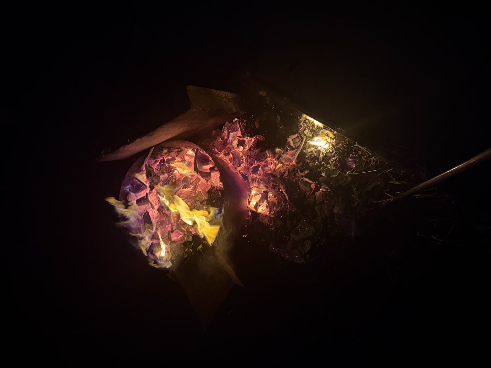 A fire bowl seen from above at night, pink and violet coals with one bright flame, framed by deep darkness.